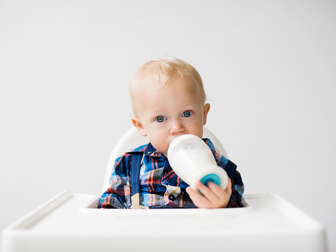 Kleiner Junge sitzt im Hochstuhl und trinkt Milch aus Milchflasche