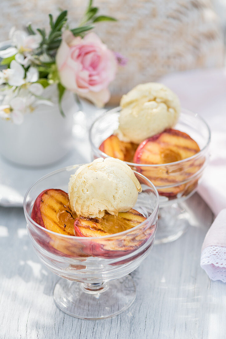 Gegrillte Pfirsiche mit Vanilleeis in Dessertschälchen auf sommerlichem Tisch im Freien