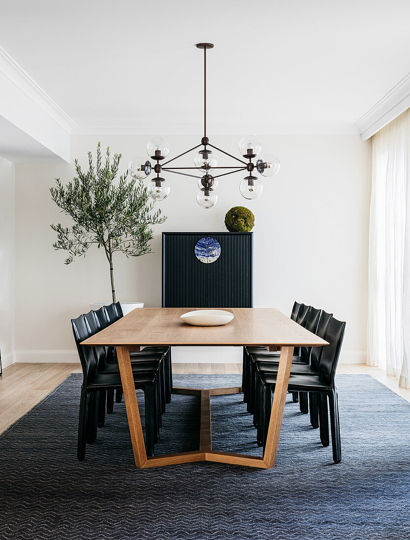 Langer Tisch mit schwarzen Stühlen unter Kronleuchter, im Hintergrund schwarzer Schrank und Olivenbäumchen