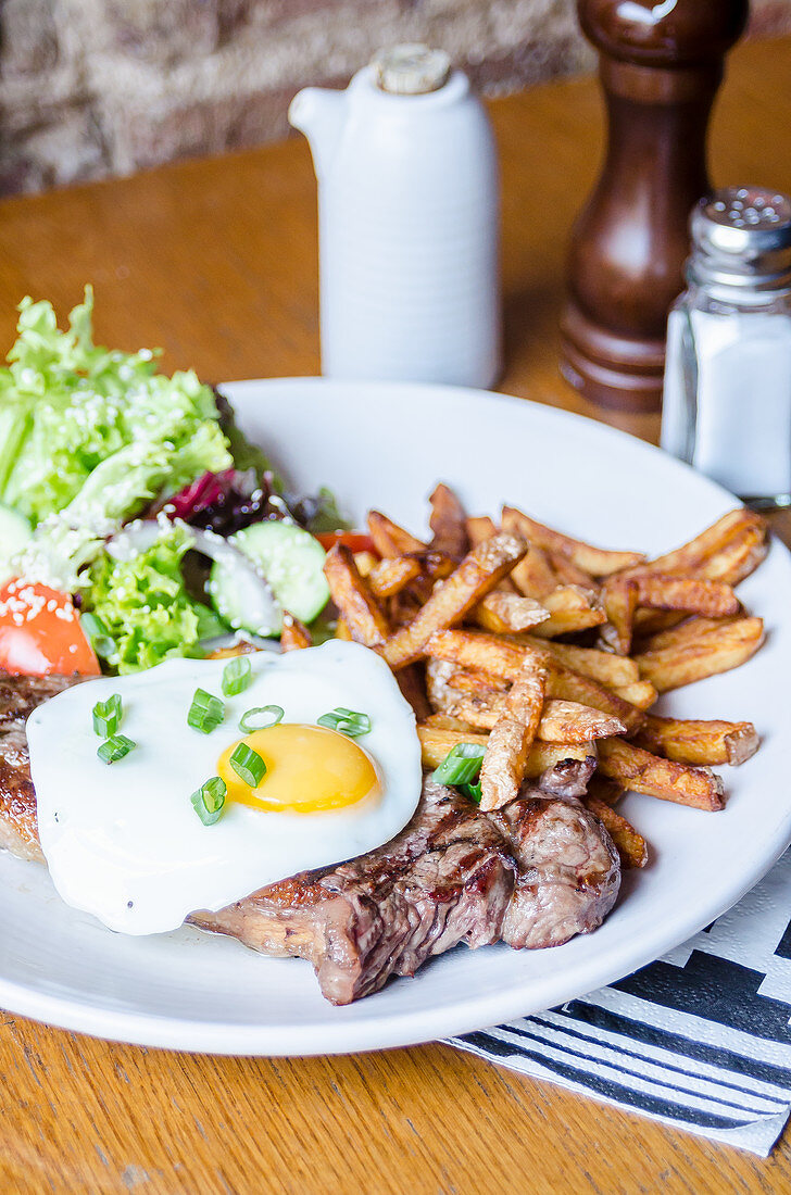 Beef rump steak topped with fried egg and chives herbs served with golden chips and fresh salad on a white plate on a wooden table