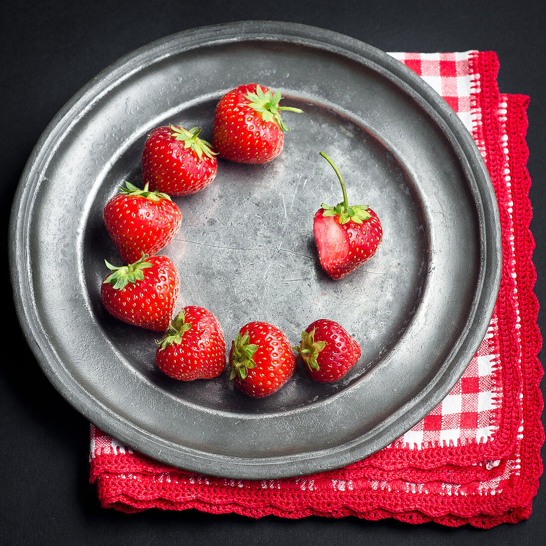 Fresh strawberries arranged circularly on a dark pewter metallic plate with a red and white napkin on a black background