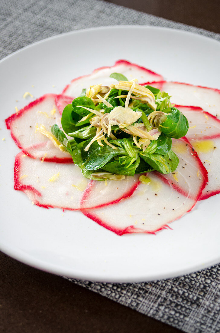 Fresh tuna carpaccio with herb salad and artichokes drizzled with olive oil on a white plate