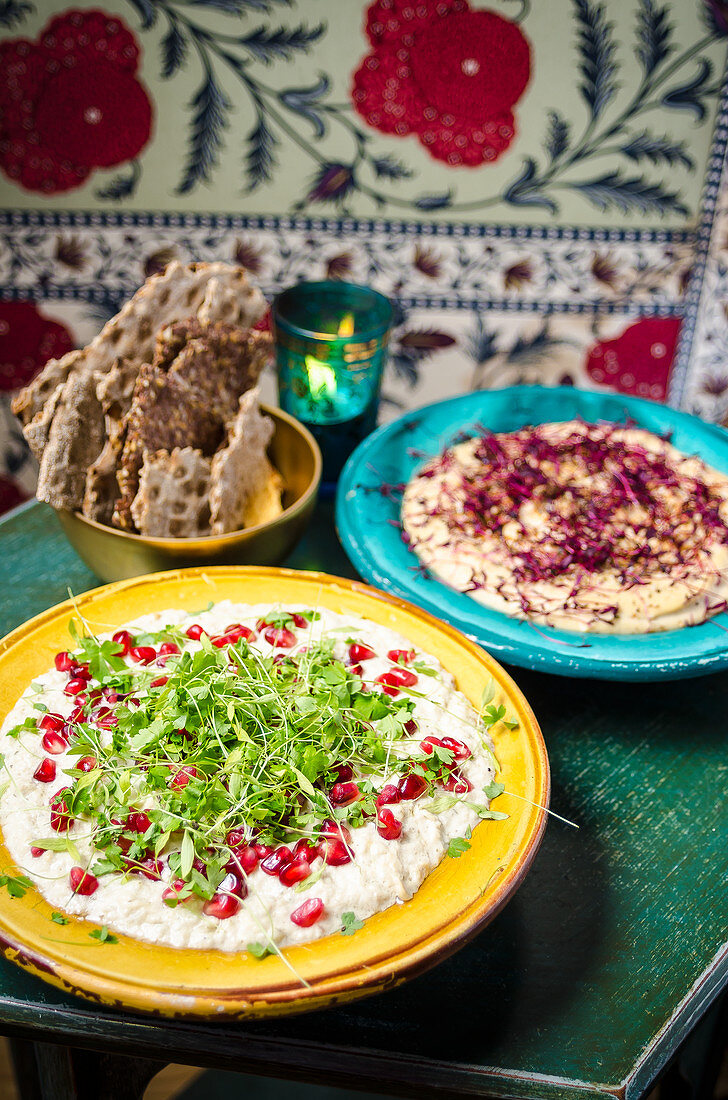 Hummus sharing plate garnished with herbs and pomegranate seeds with crispy flat bread pieces in colourful plates and background