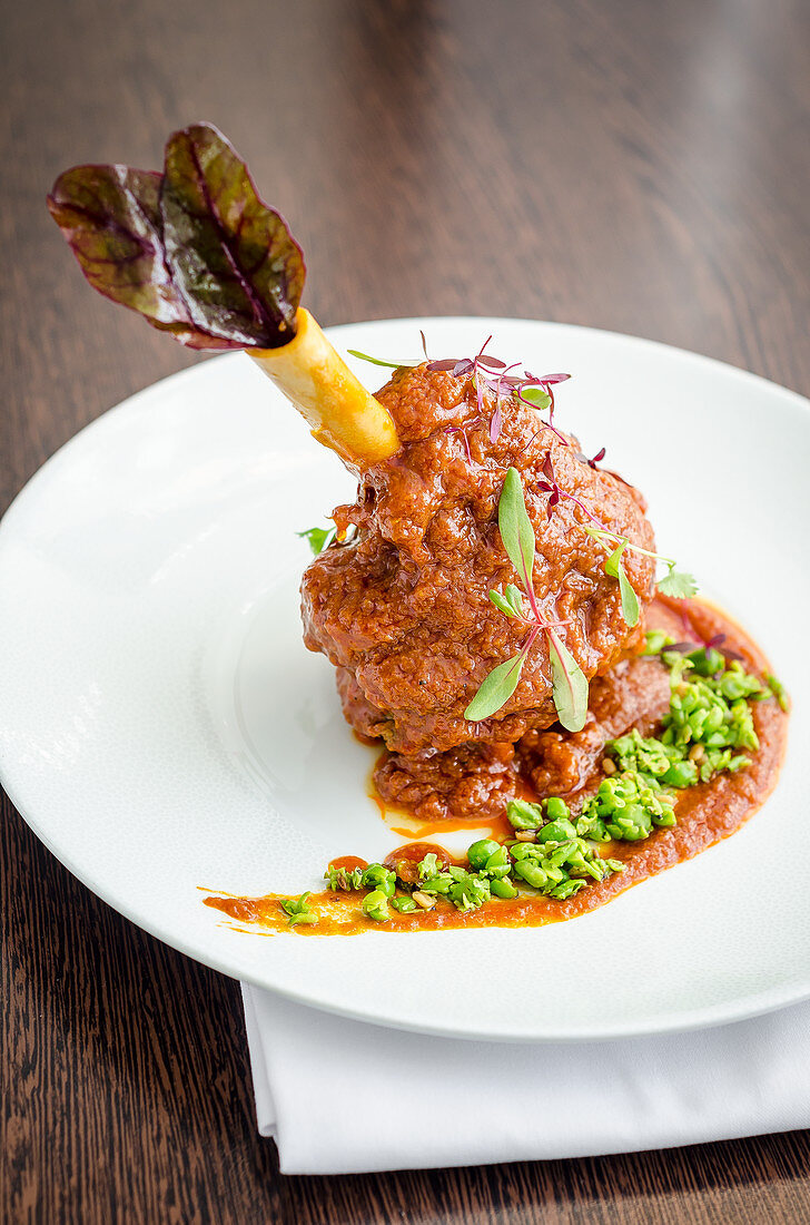 Slow cooked lamb shank in a rich tomato, yogurt, turmeric and peas sauce on a white plate and wooden background