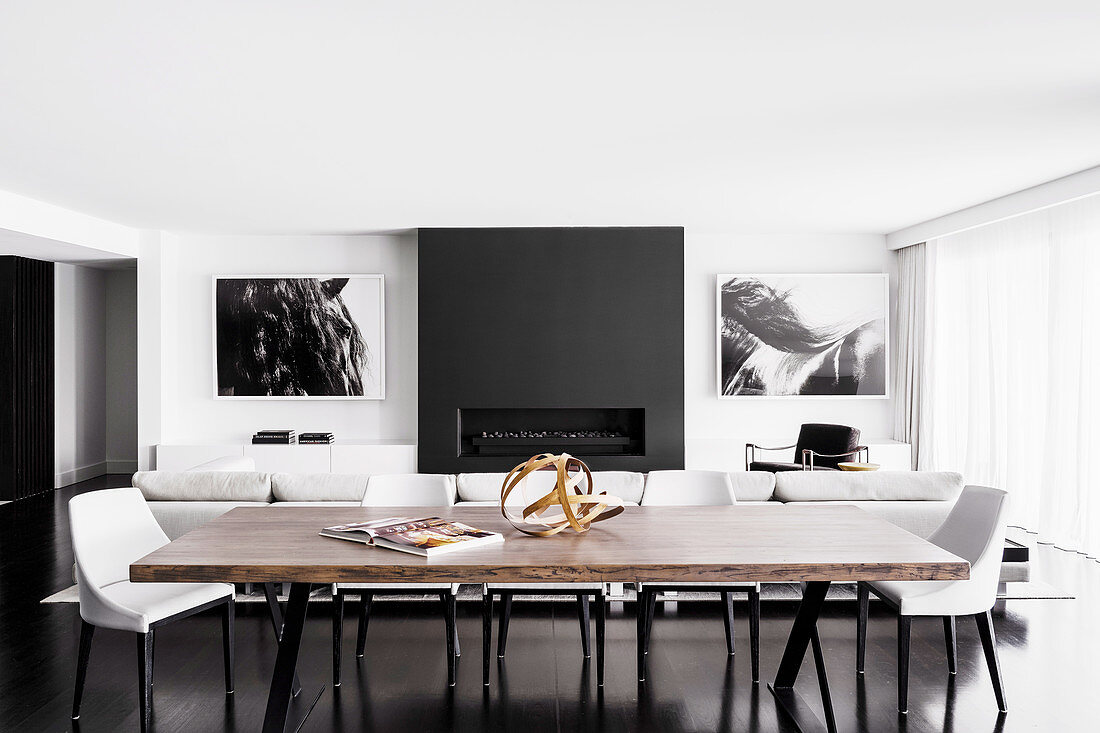 Long dining table with wooden top and designer chairs in front of white sofa set and fireplace in open living room