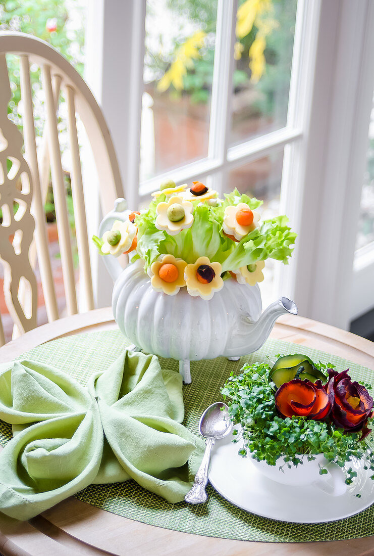 Bouquet of salad leaves and flowers made with cheese, carrots and avocado in a teapot, cress and carrots in a tea cup on a green tablemat