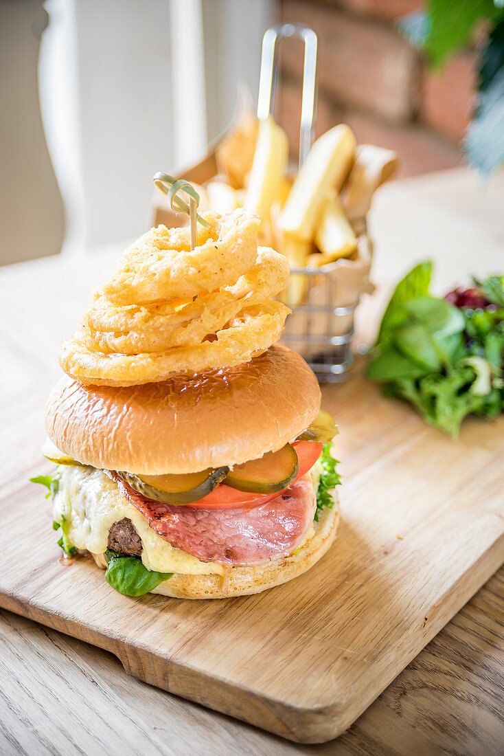 Bacon cheese beef burger with gherkins, pickle, tomato, fresh salad, bacon, cheese and fried onion rings ona wooden board with chips and salad
