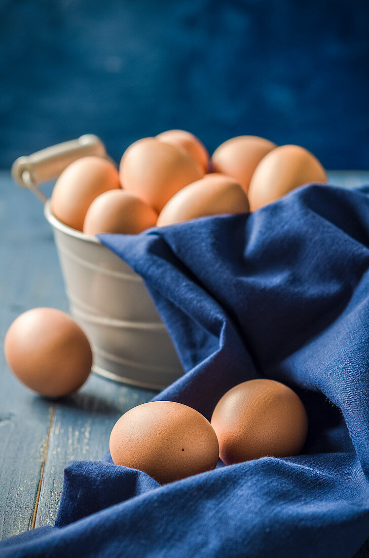 Fresh eggs in a rustic metal basket and blue drape on a blue wooden table and a blue background