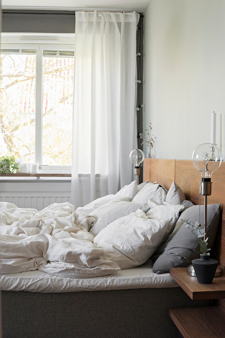 Ungemachtes Doppelbett mit Holzkopfteil, Glühfadenlampe auf dem Nachttisch