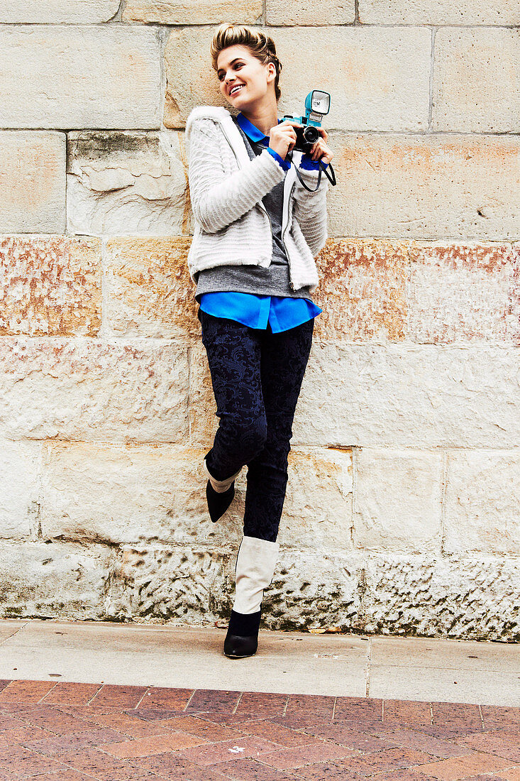 A blonde woman wearing a fleece jacket and jeans with a brocade pattern holding a camera