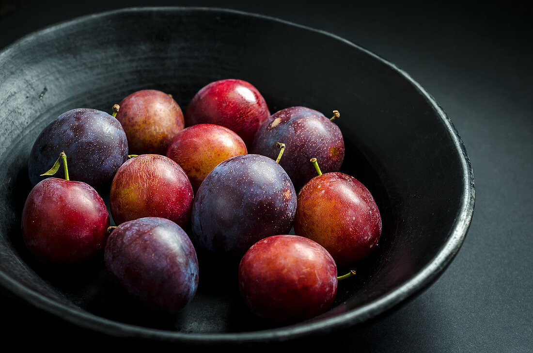 bowl of fresh purple and red prunes and plums in a black fruit bowl on a black background