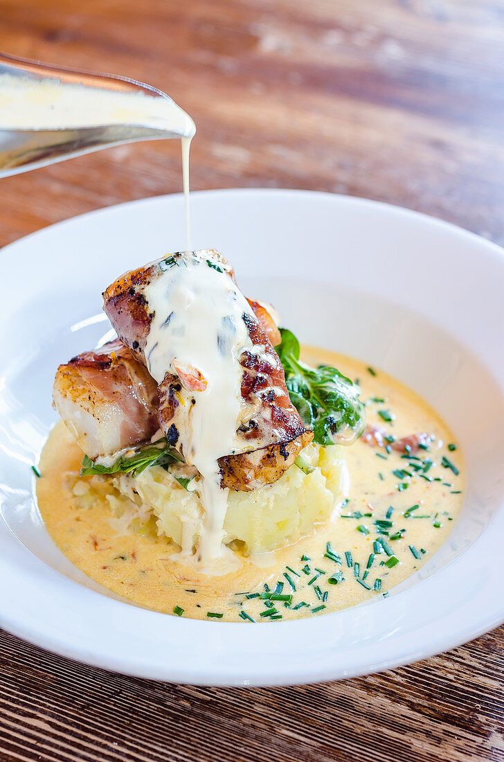 Sea bass fish fillets wrapped in bacon on a potato mash with spinach and chives with a pour of white wine sauce