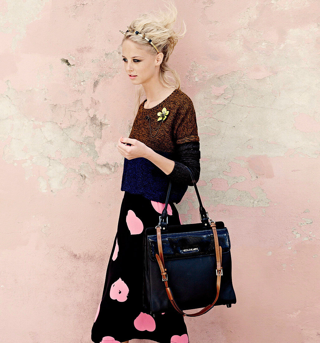 A young blonde woman wearing a jumper, a black skirt with pink hearts with a handbag and a hair band