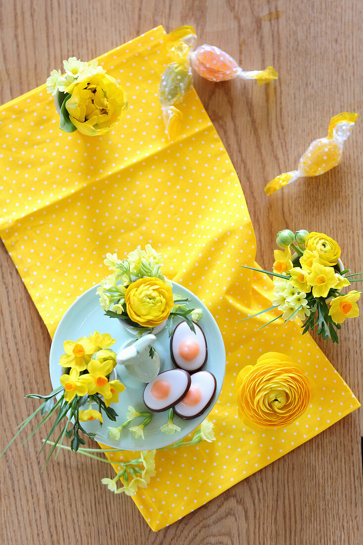 Egg-shaped sweets on bunny cake stand and spring flower in egg cups on yellow cloth
