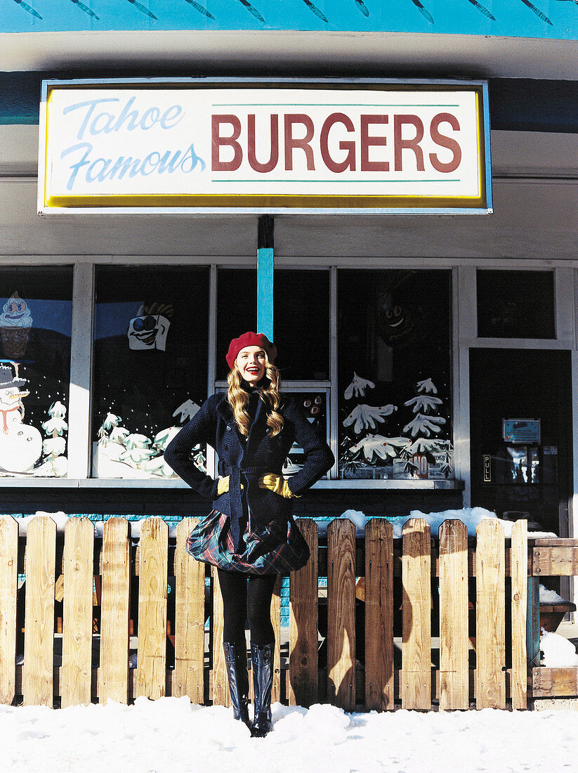 A young woman outside a burger restaurant wearing a dark jacket, a skirt, tights and boots