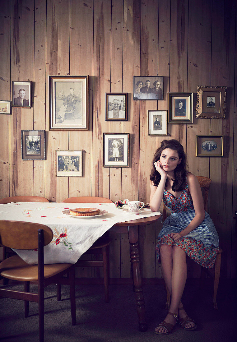 A dark-haired woman in a wooden cabin wearing a floral dress and an apron