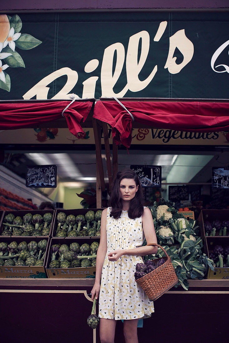 A dark-haired woman outside a vegetable shop wearing a summer dress