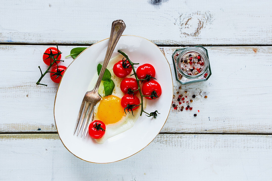 Healthy breakfast set with plate of fried egg, roasted cherry tomatoes, basil and spices on white wooden backdrop