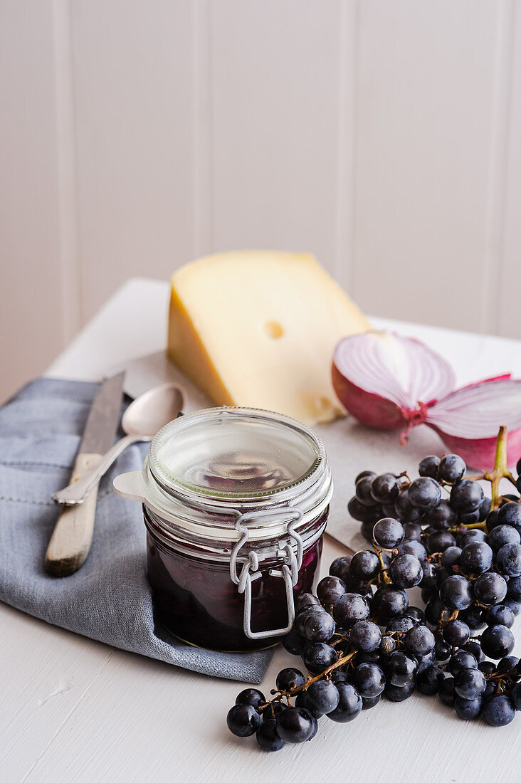 A jar of red grape chutney surrounded by cheese, onions and grapes
