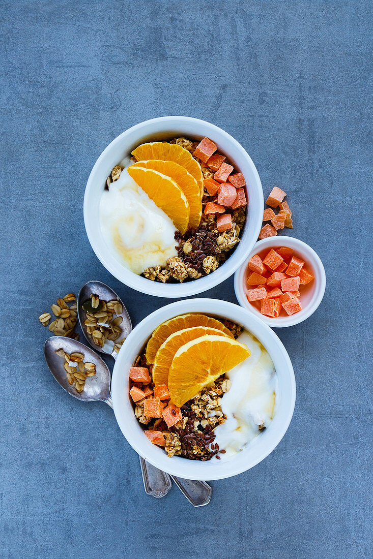 Vegetarian breakfast yogurt bowls with tasty granola, orange, dried fruit, nuts and seeds over grey background, top view