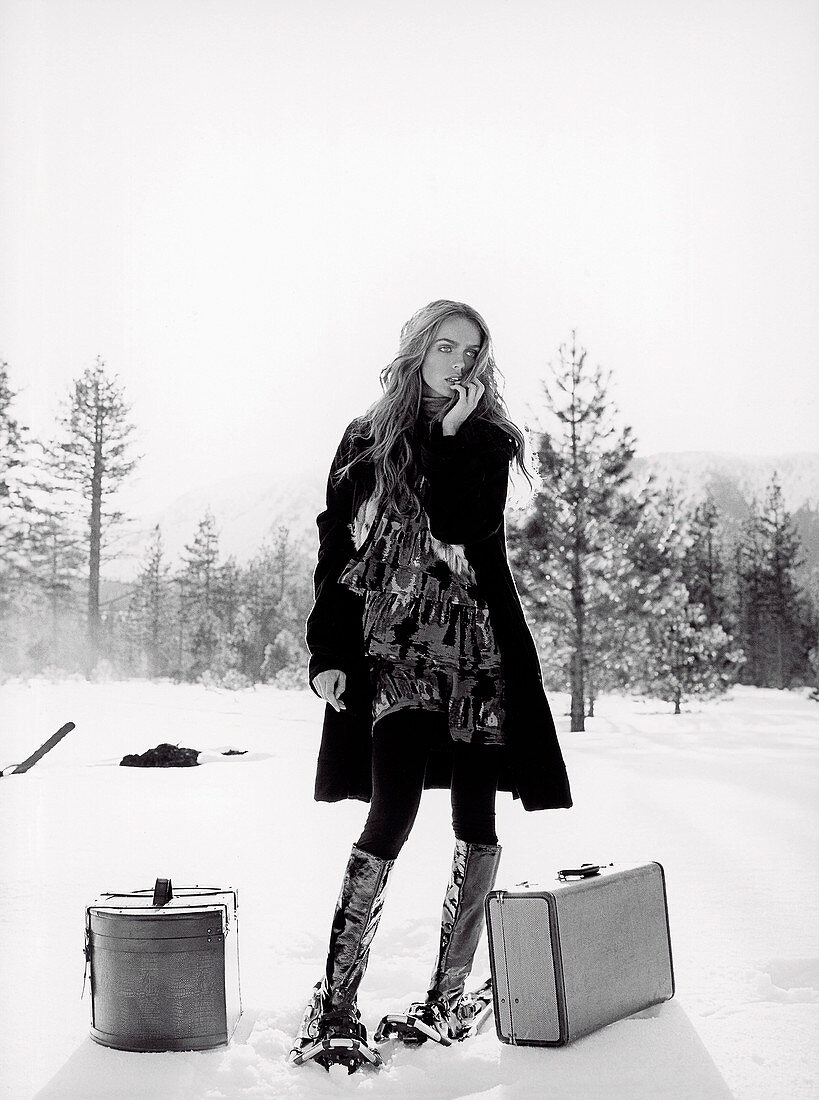A young woman in the snow with suitcases wearing a dress and a dark coat (black-and-white shot)