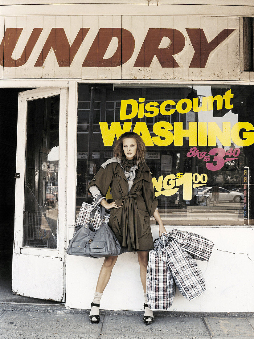 Junge Frau im Trenchcoat mit großen Taschen vor Waschsalon
