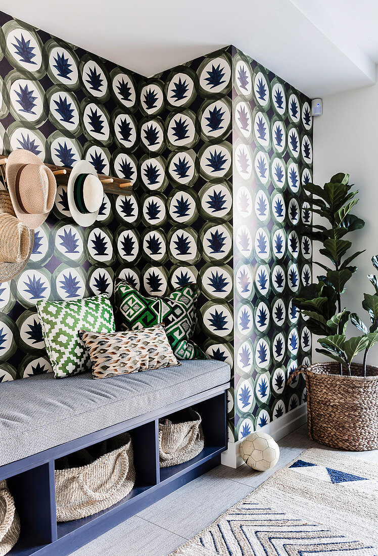 Bench with upholstery and cushions, coat rack with hats and houseplant in front of wallpapered wall