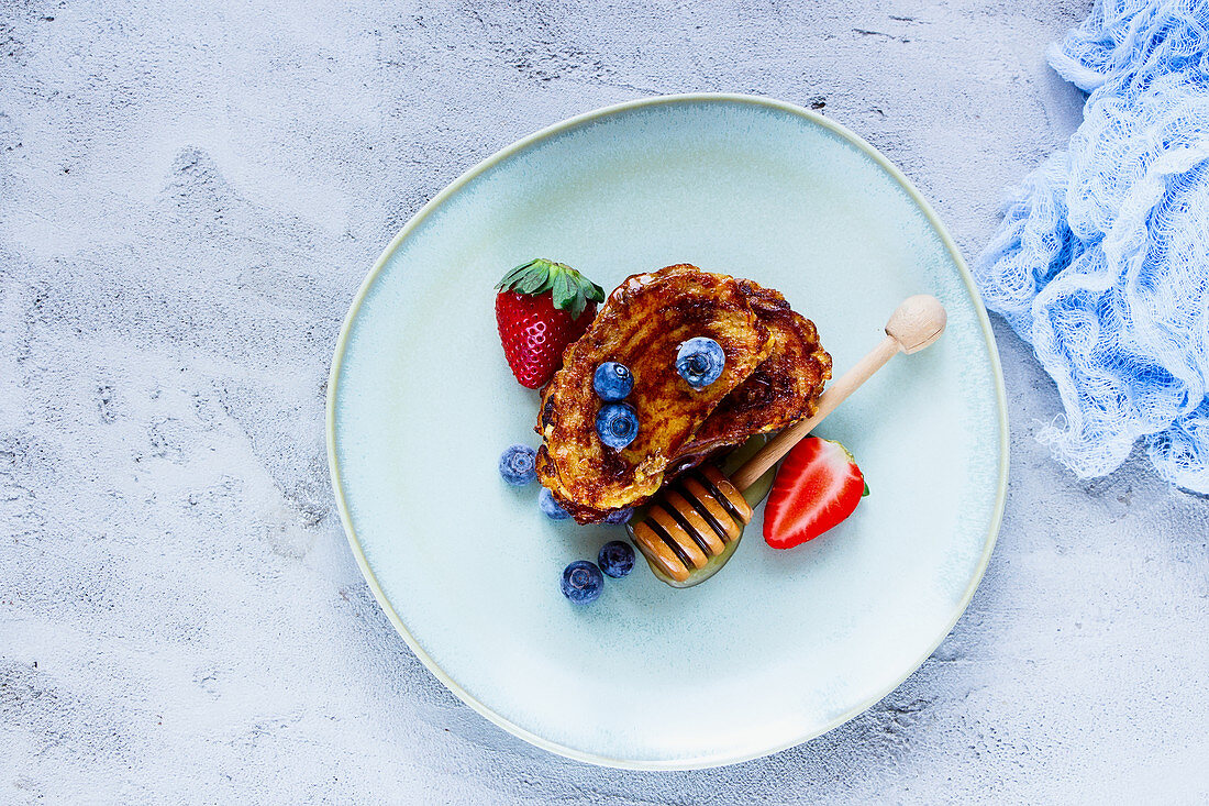 Homemade cinnamon french toasts with blueberries, strawberries and honey in ceramic plate for tasty breakfast on concrete textured background