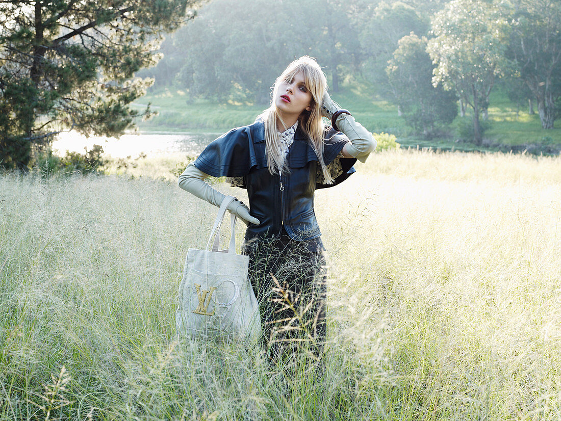 A young blonde woman standing in a meadow wearing a leather jacket and a skirt