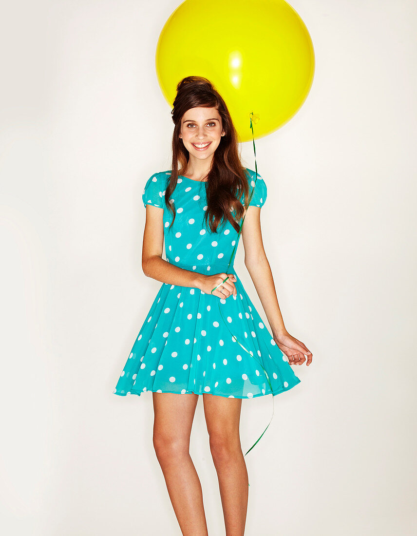 A young dark-haired woman with a yellow balloon wearing a turquoise, polka dot dress