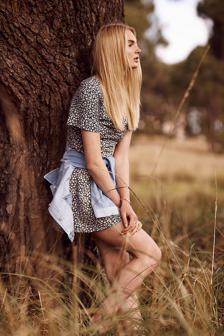 A young blonde woman by a tree wearing a printed dress with a denim jacket around her waist