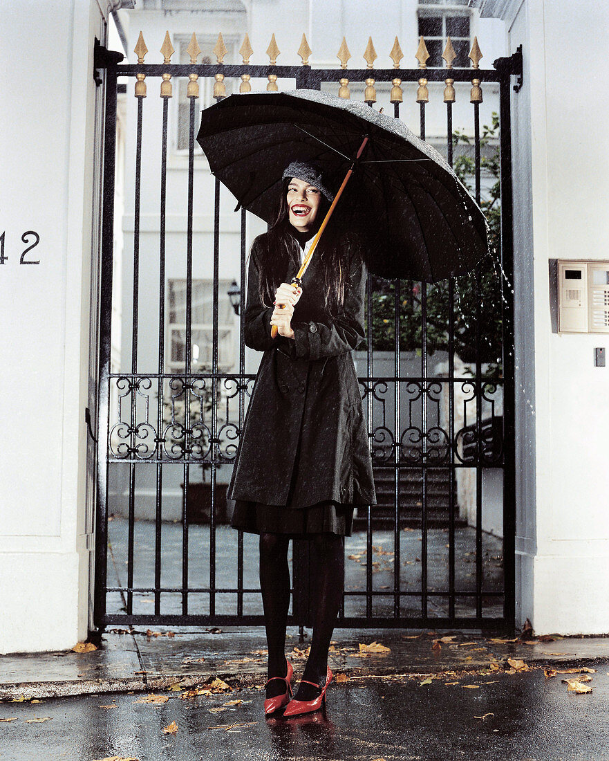 A young woman wearing a black trench coat with a black umbrella standing outside a wrought-iron gate