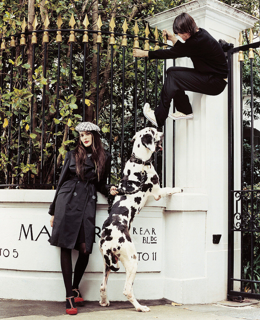 Junge Frau in schwarzem Trenchcoat mit Dalmatiner, junger Mann klettert am Zaun hoch