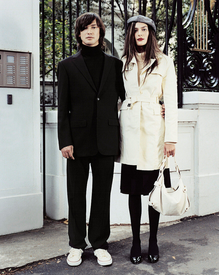 A dark-haired woman wearing a beret and a light trench coat with a man in a black outfit