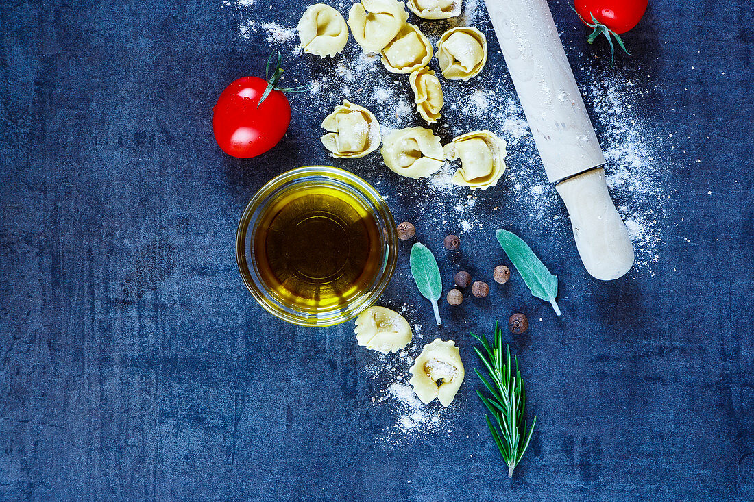 Food background with homemade Italian pasta tortellini, tomatoes, flour, fresh herbs and olive oil on dark vintage texture