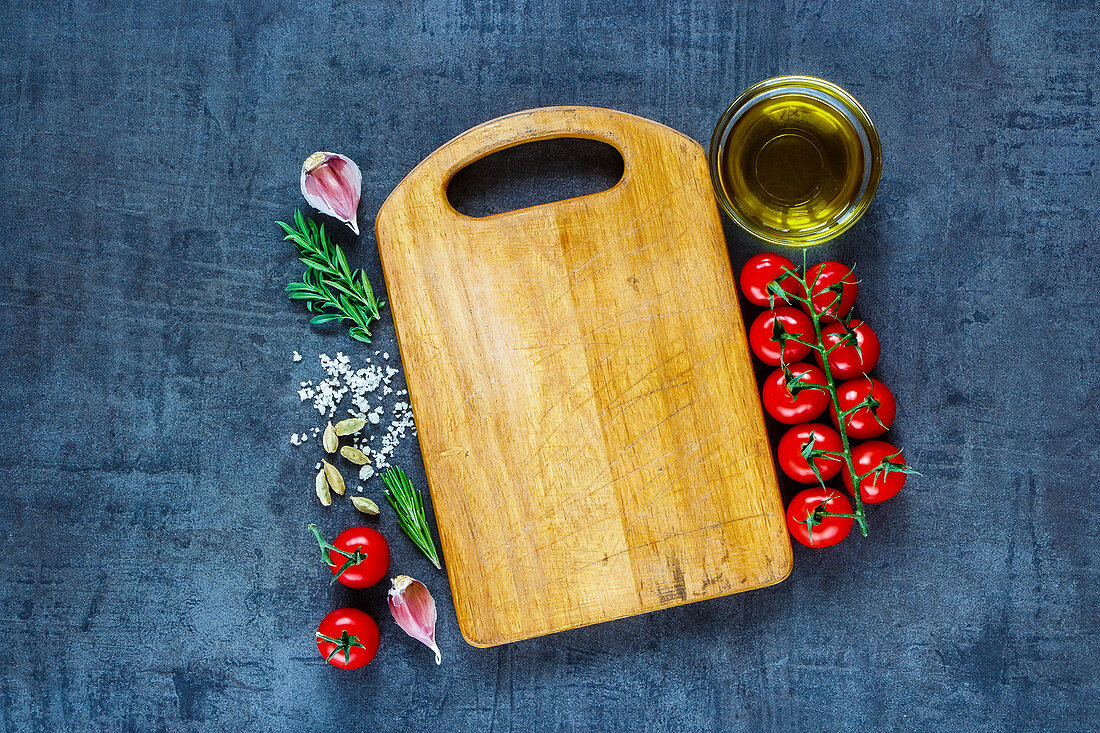 Holzbrett umgeben von Tomaten, Olivenöl, Knoblauch, Rosmarin und Salz