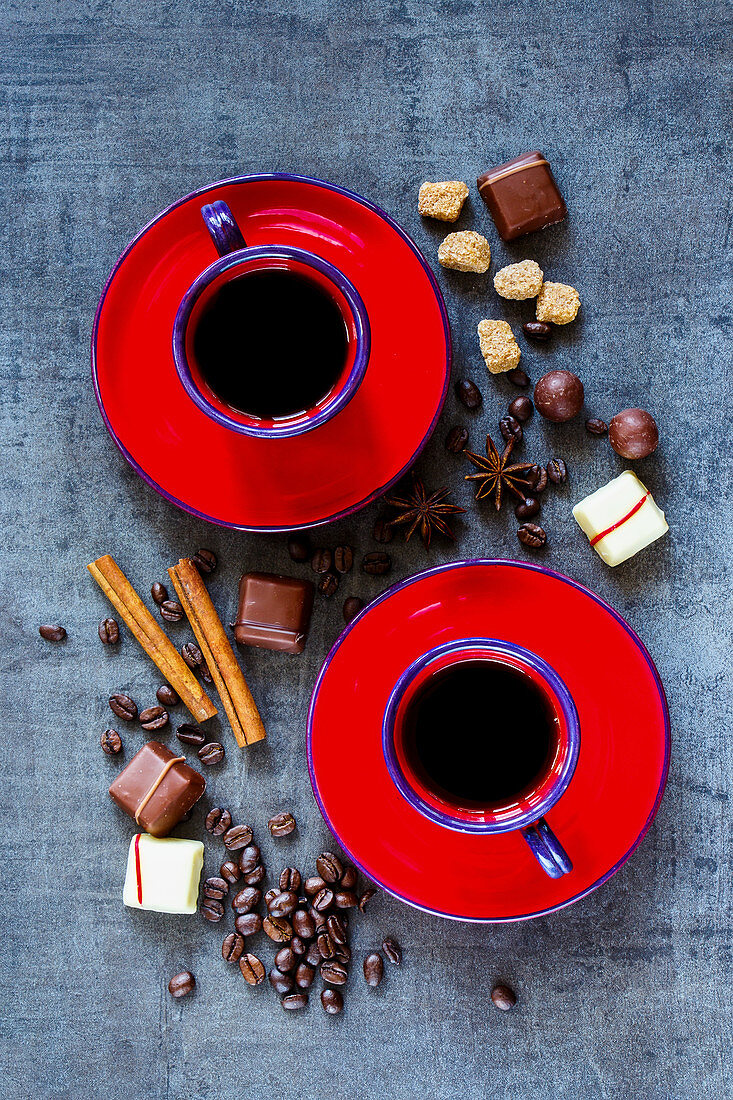 Kaffeetassen mit roten Untertassen, Gewürzen und Schokolade auf grauem Untergrund