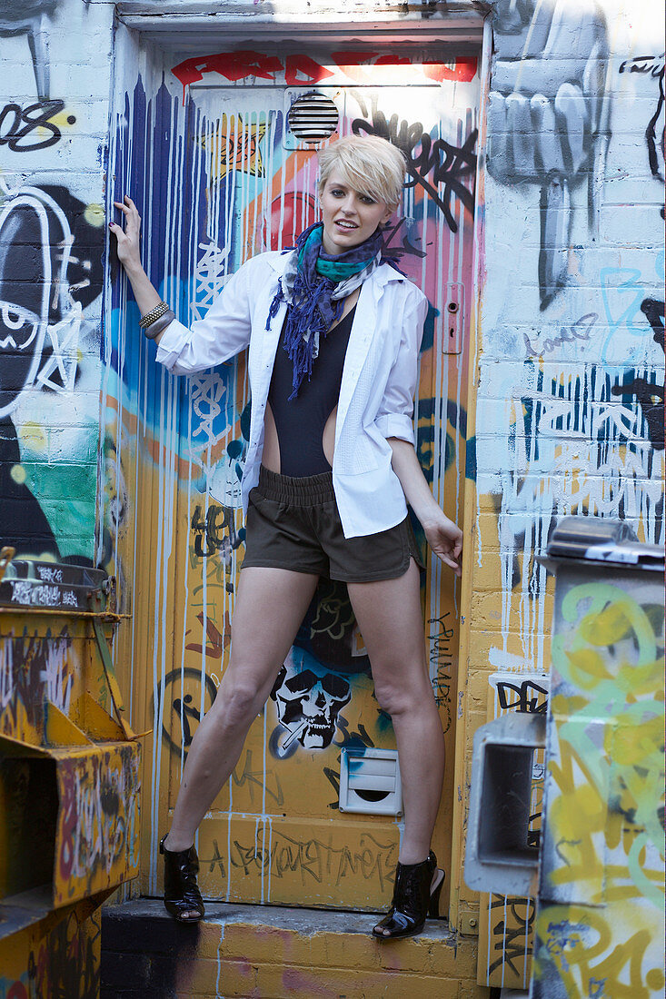 A blonde woman wearing a white blazer and shorts standing by a graffitied door