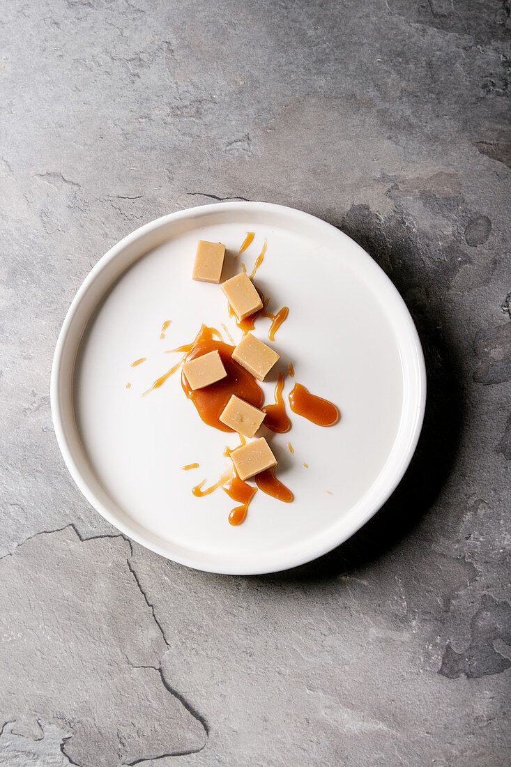 Salted caramel fudge candy with caramel sauce in white plate over grey texture background
