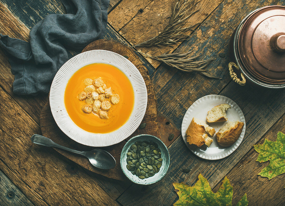 Flat-lay of fall warming pumpkin cream soup with croutons and seeds on board over rustic wooden background