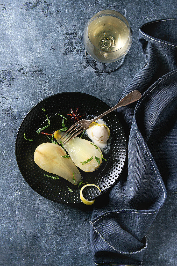 Traditional dessert sliced poached pears in white wine served in black plate with syrup, ice cream, lemon zest and glass of white wine