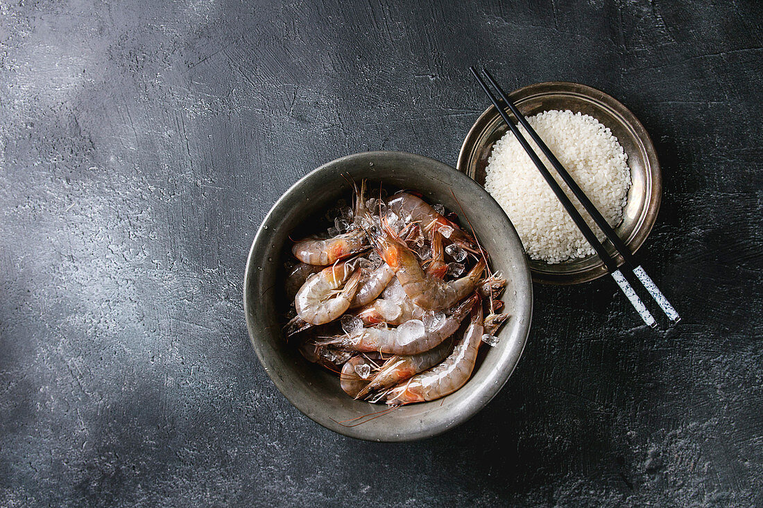 Raw whole fresh uncooked prawns shrimps and plate of raw rice and chopsticks over black texture background