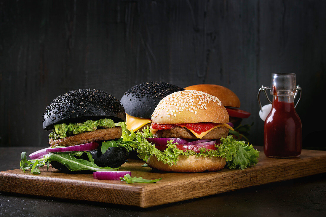 Homemade burgers in black and white buns with avocado, tomato sauce, lettuce, arugula, cheese, onion on wood serving board