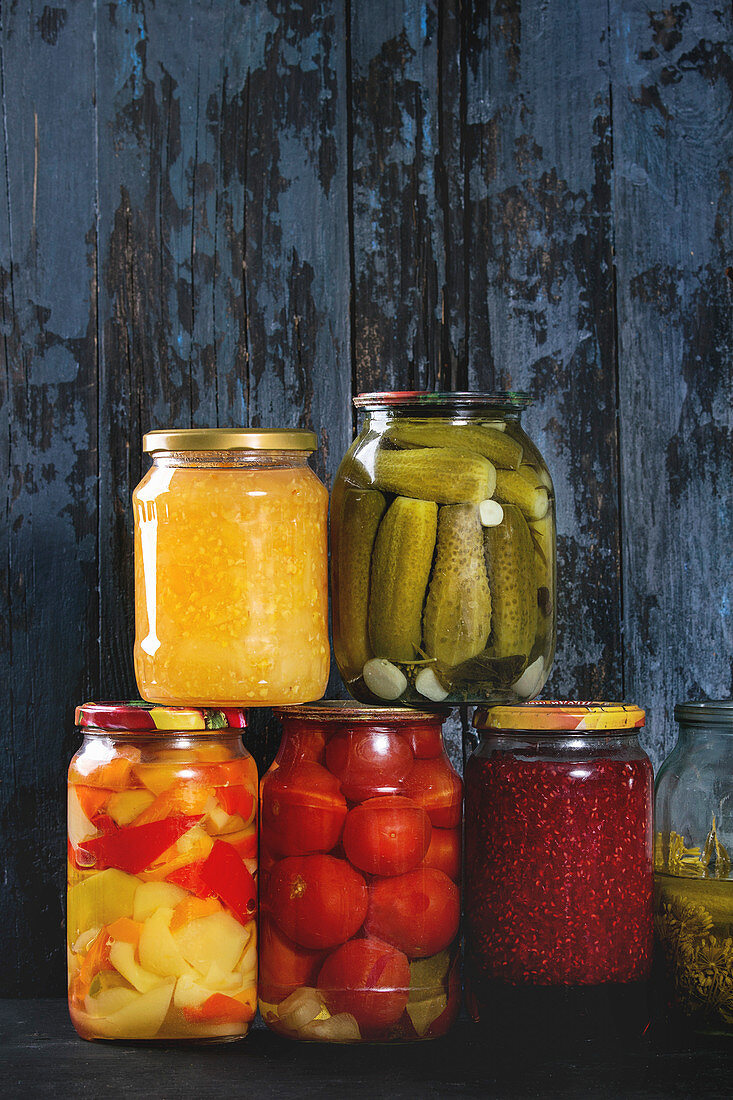 Variety glass jars of homemade pickled or fermented vegetables and jams in row with old dark blue wooden plank background