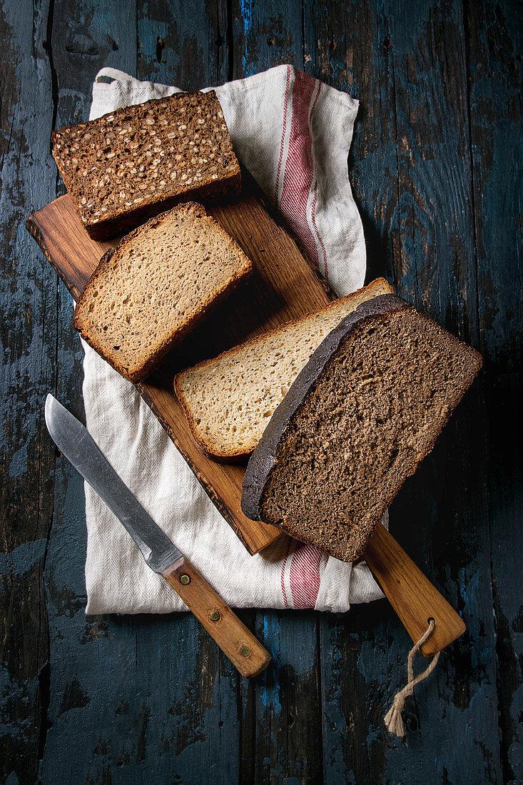 Variety loaves of sliced homemade rye bread whole grain and seeds on wooden cutting board with kitchen towel and knife
