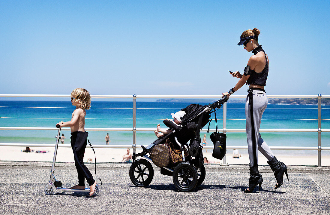 A young woman with a baby in a buggy and a young child on a scooter by the beach