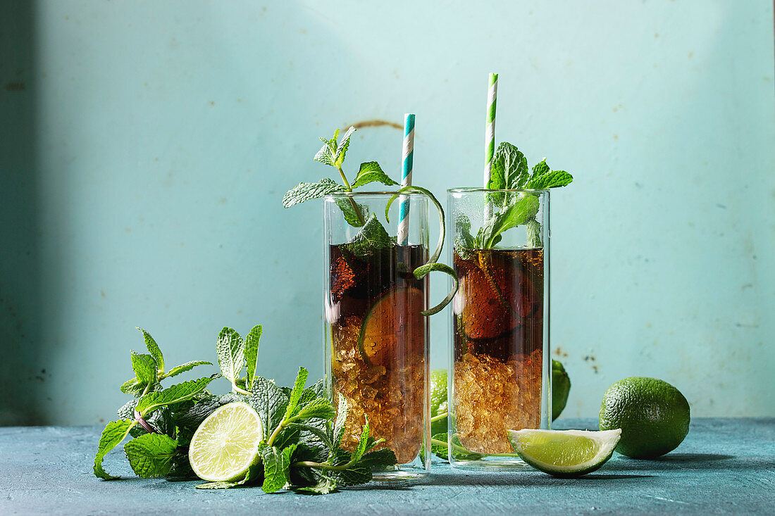 Two Glasses of classic Cuba libre cocktail with lime, mint, crushed ice and cola, served with ingredients above and retro cocktail tubes
