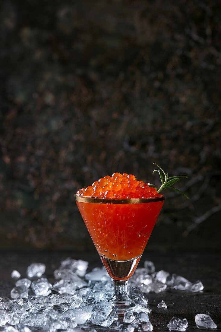 Glass of red caviar decorated by fresh rosemary on crushed ice over dark background