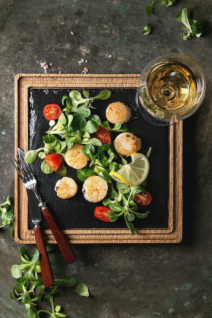 Fried scallops with lemon, cherry tomatoes and green salad served on wooden black slate serving board with cutlery and glass of white wine over old dark metal background