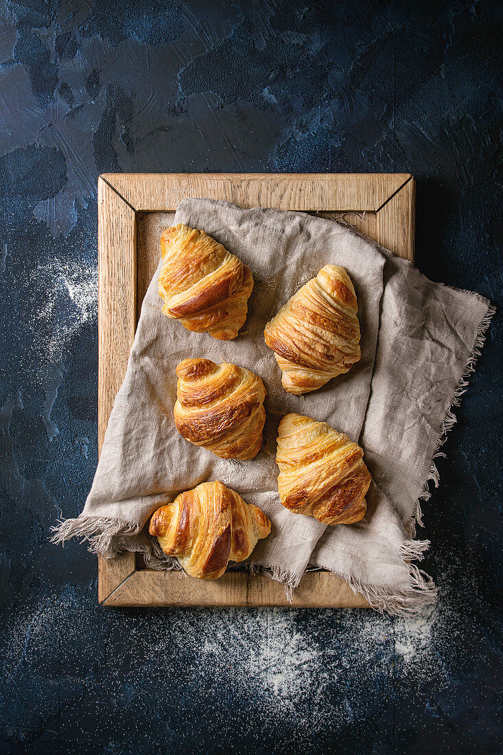 Fresh baked traditional croissants on linen textile wooden tray over dark blue texture background